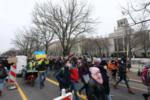Eine große Protestdemonstration mit Menschen, die eine Straße entlanggehen, einige halten Schilder und fahren Fahrräder, mit Bäumen und einem klaren blauen Himmel im Hintergrund.
