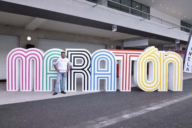 Ein Mann steht an der Startlinie eines Marathons vor einem großen "Marathon"-Schild, mit einem Banner rechts daneben und einem Gebäude mit Lichtern, Geländern und Läden im Hintergrund.