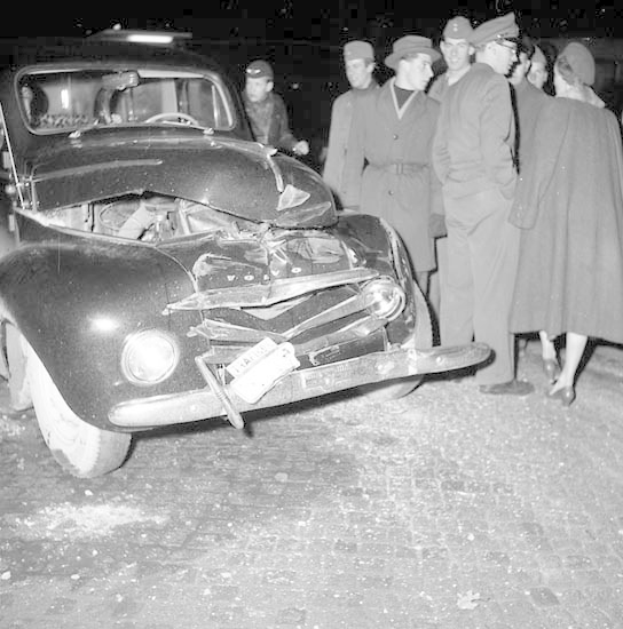 Schwarz-weißes Bild eines schwerbeschädigten Autos am Straßenrand mit einer Gruppe von Menschen, die es inspizieren.