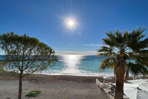 Eine sonnige Strandszene mit Palmen, Sonnenschirmen und üppiger Vegetation, die vor einem hellblauen Himmel mit der Sonne im Hintergrund liegt, geeignet für eine Ferienwohnung an der französischen Riviera.