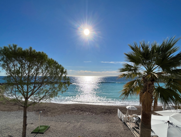 Eine sonnige Strandszene mit Palmen, Sonnenschirmen und üppiger Vegetation, die vor einem hellblauen Himmel mit der Sonne im Hintergrund liegt, geeignet für eine Ferienwohnung an der französischen Riviera.
