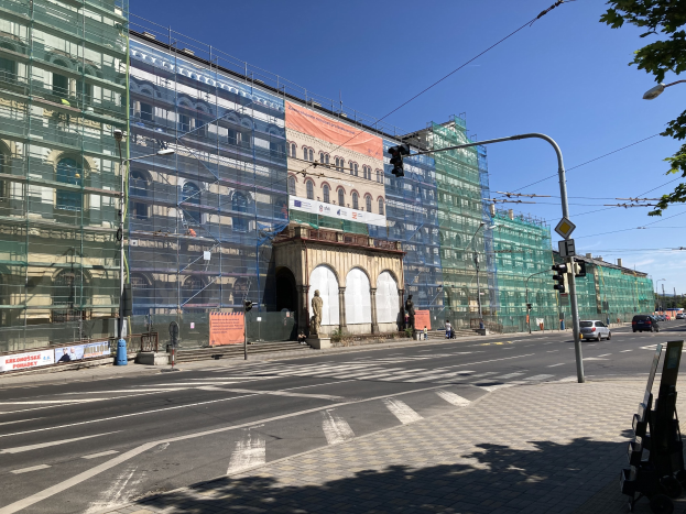 Großer mehrstöckiger Bau im Bau in Berlin, umgeben von Straßeninfrastruktur, Fahrzeugen, Fußgängern und Bäumen bei bewölktem Himmel.