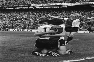 Drei Fußballspieler feiern ein Tor auf dem Boden, mit jubelnder Menge und schwenkenden Fahnen im Hintergrund, in Schwarz-Weiß aufgenommen.