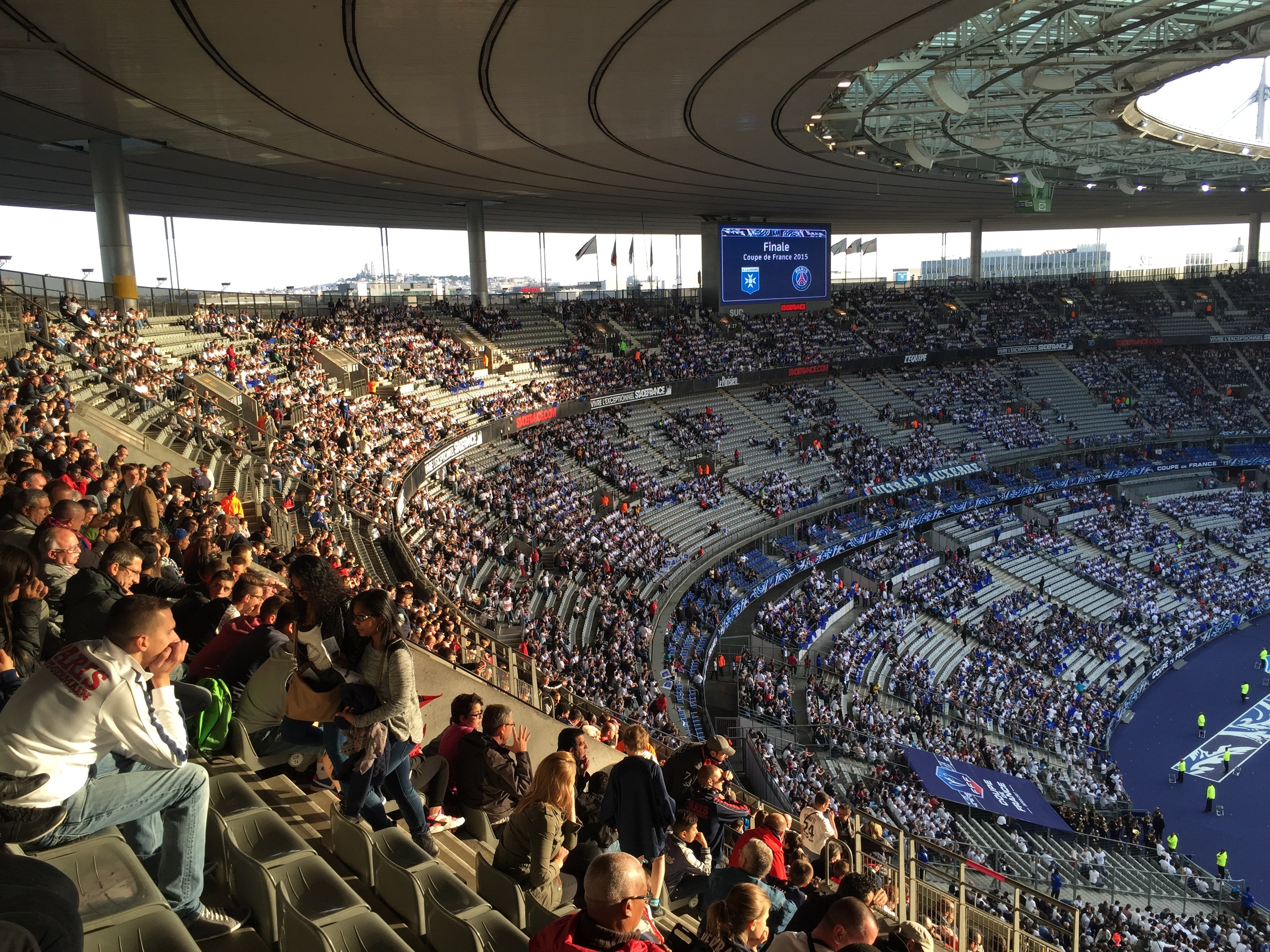 Großes Publikum in einem Stadion bei einem Fürballspiel, mit einer Bühne auf der rechten Seite, Fahnen, Masten, einem Bildschirm und der Allianz Arena in München, Deutschland im Hintergrund.