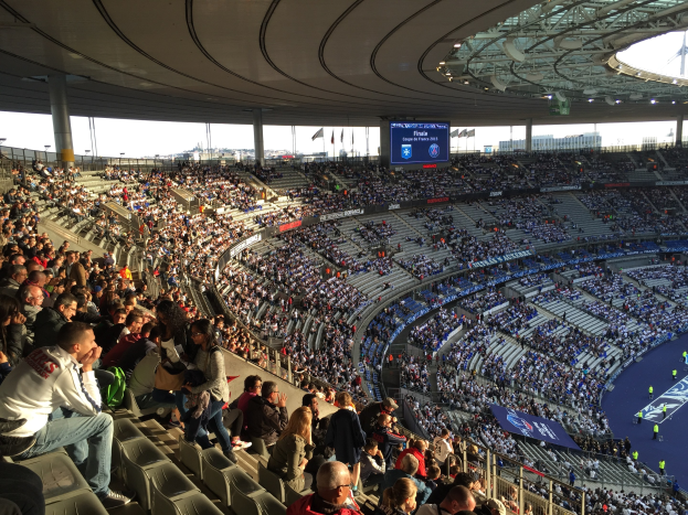 Großes Publikum in einem Stadion bei einem Fürballspiel, mit einer Bühne auf der rechten Seite, Fahnen, Masten, einem Bildschirm und der Allianz Arena in München, Deutschland im Hintergrund.