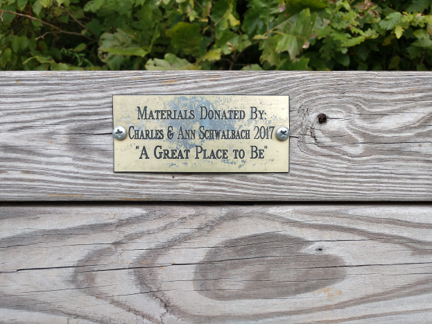 Eine hölzerne Bank mit einer Tafel, auf der steht: "Materialien gespendet von Charles & Ann Schwabach 2017: Ein toller Ort zu sein" umgeben von üppigen grünen Pflanzen.