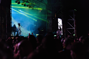 Eine Menge schaut einer Band zu, die auf einer hell beleuchteten Bühne mit Instrumenten, Mikrofonen und einem Banner spielt, vor einem dunklen Hintergrund.