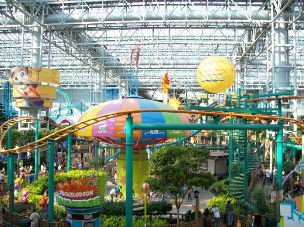 Ein großes Indoor-Wasserpark mit einer Achterbahn, umgeben von Menschen, Bäumen, Pflanzen und Deckenbeleuchtung, die eine lebendige Atmosphäre schaffen.