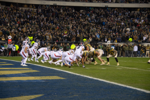 Ein Fußballspiel im Gange mit Spielern in Helmen auf dem Feld, Zuschauern, Geländern und Tafeln mit Text im Hintergrund.