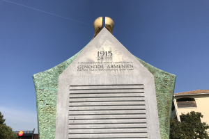 Ein steinernes Denkmal mit einer Uhr darauf, umgeben von Bäumen und Gebäuden, mit Text, der den 24. Jahrestag des Völkermords in Armenien erinnert.
