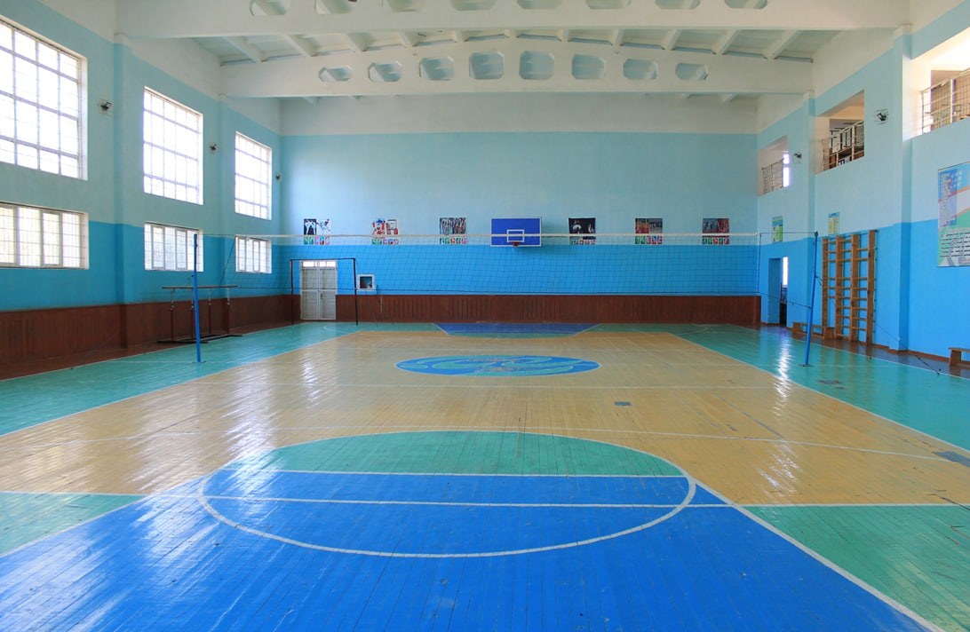 Große Turnhalle mit blau-weißem Boden, Basketballfeld, Netz, Stangen, Tür, Fenstern, Plakaten, Geländer und Dach.