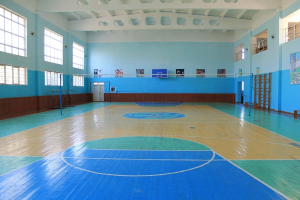 Große Turnhalle mit blau-weißem Boden, Basketballfeld, Netz, Stangen, Tür, Fenstern, Plakaten, Geländer und Dach.