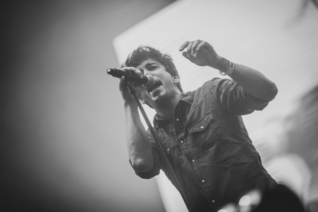 Schwarz-weiß-Foto eines Mannes, der in ein Mikrofon auf der Bühne singt, mit einem unscharfen menschlichen Gesicht im Hintergrund.