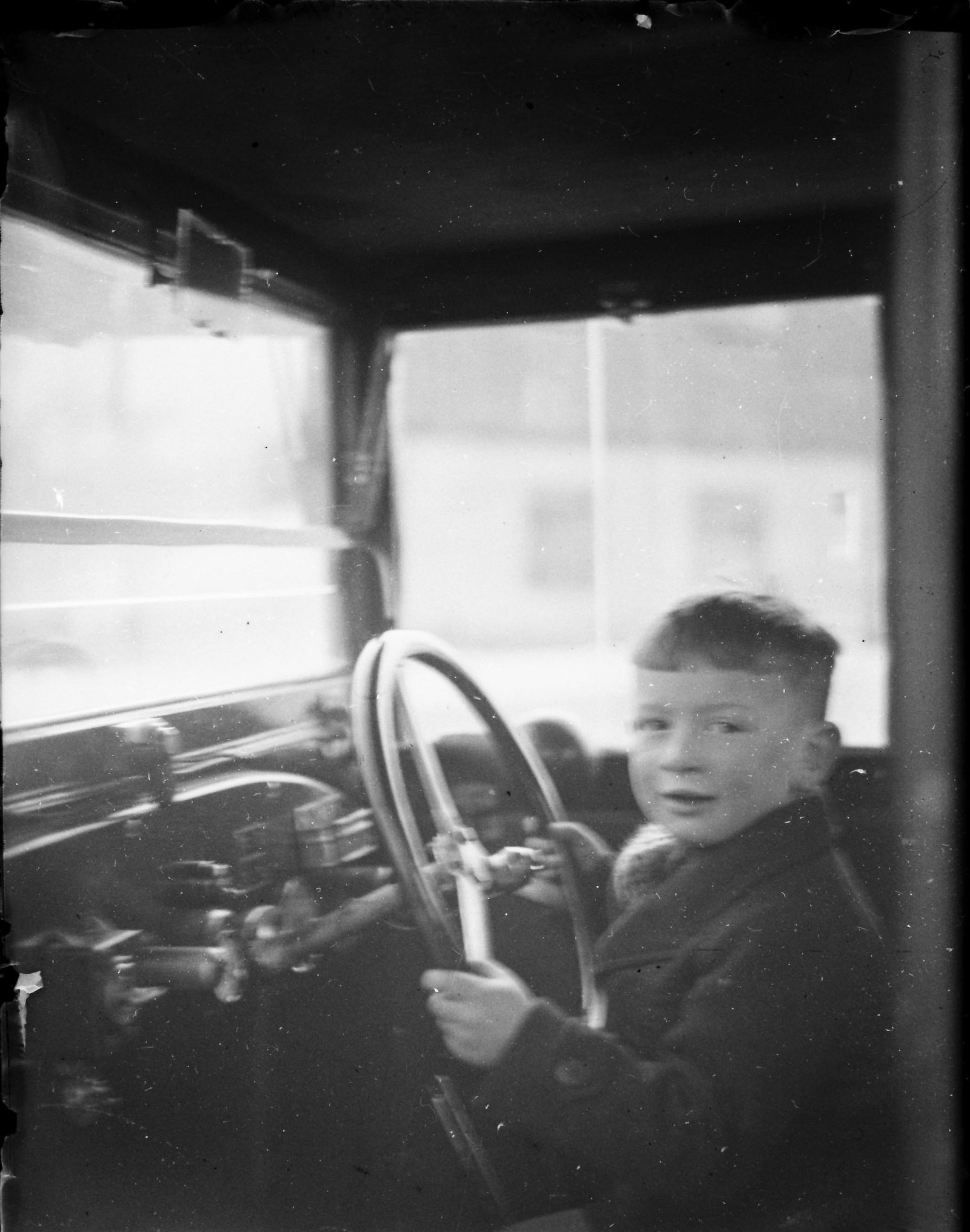 Ein junger Junge in Schwarz-Weiß sitzt im Fahrersitz eines Autos und hält das Lenkrad konzentriert fest.