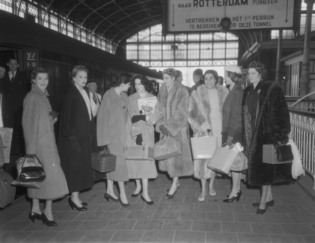 Schwarz-weißes Bild von Frauen, die an einem Bahnhof mit Taschen stehen, neben einem Zug und unter einer Decke mit Geländern.