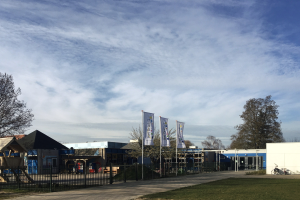 Großes Gebäude mit Metallzaun, Rasenfläche, Weg, Bäumen, Pfählen, Bannern, Fahrrad, nahen Häusern und bewölktem Himmel, identifiziert als neuer Spielbereich für Kinder an der Schule.