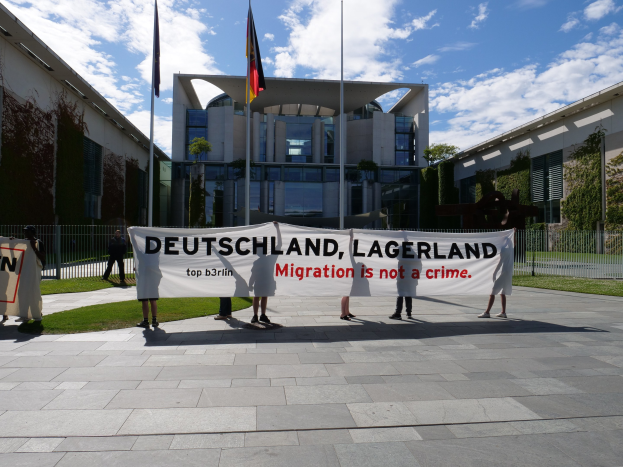 Gruppe von Menschen mit einem Banner, auf dem "Deutschland, Lagerland Migration ist kein Verbrechen" steht, vor einem Gebäude mit Fenstern, umgeben von Gras, einem Metallzaun, Pflanzen, Bäumen und Fahnen mit Stangen unter einem bewölkten Himmel.