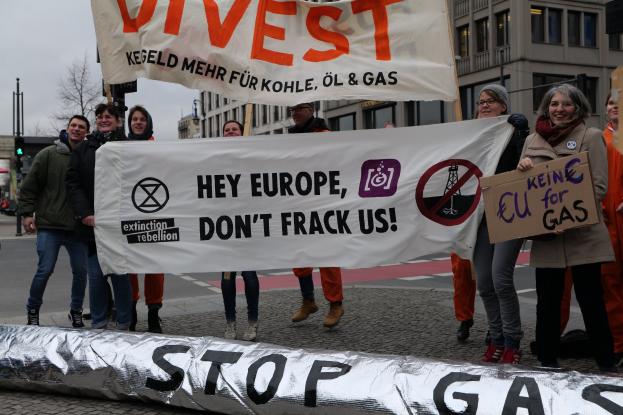 Gruppe von Menschen, die auf einer Straße protestieren und ein "Stoppt Gas"-Schild gegen die Europäische Union halten, mit Gebäuden, Bäumen und Verkehrszeichen im Hintergrund unter einem klaren blauen Himmel.