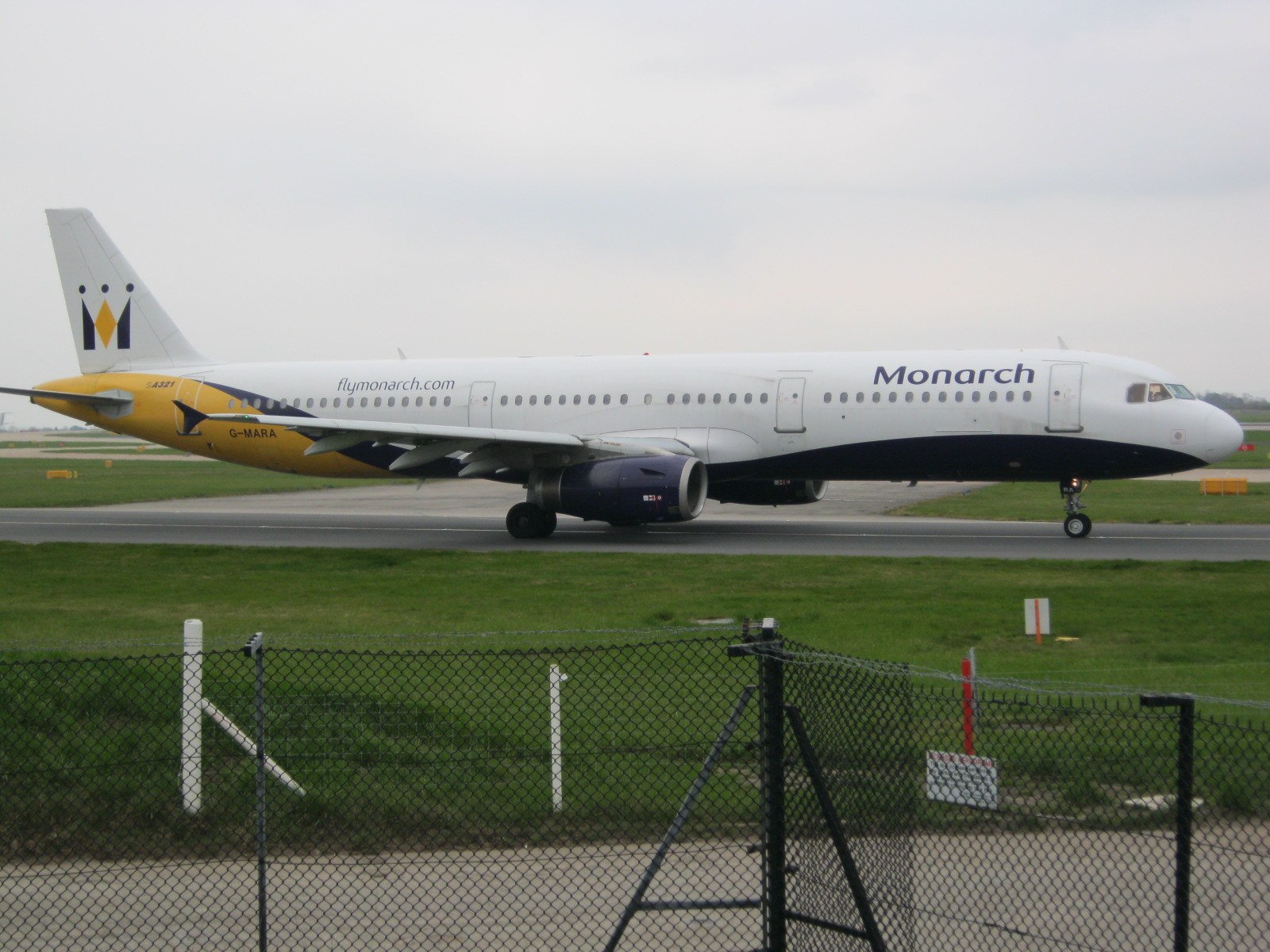 Eine Monarch Airlines Airbus A320-200 auf der Rollbahn, umgeben von Gras und einem Zaun mit Pfählen und Brettern, mit Bäumen und einem klaren blauen Himmel im Hintergrund.