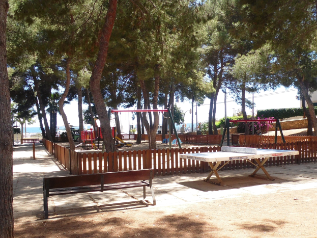 Eine Parkszene mit einem zentralen Tischtennis-Tisch, umgeben von Bänken, einem Zaun, Schaukeln, Pfählen, Pflanzen und Bäumen, mit einem Wasserbecken und einem klaren blauen Himmel im Hintergrund.