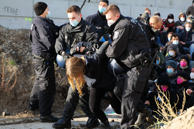 Gruppe maskierter Personen in Mützen, einige bewaffnet, die eine Frau umgeben, die von der Polizei eskortiert wird, mit städtischer Vegetation und einem protestbezogenen Wandtext im Hintergrund.