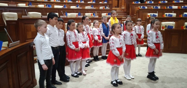Eine Gruppe von Kindern in weißen und roten Kleidern steht in einem Raum mit einem Podium, einem Mikrofon, Flaggen, Stühlen und Tischen im Hintergrund.