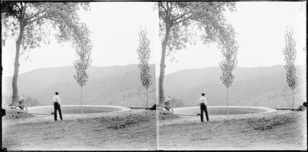 Ein Schwarz-Weiß-Foto eines Mannes, der vor einem Teich mit Bäumen, Hügeln und einem klaren Himmel steht, umgeben von üppiger Vegetation.