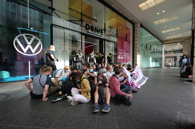 Eine Gruppe von Menschen sitzt vor einer Volkswagen-Niederlassung auf dem Boden, einige halten ein Transparent, während sie vor dem Glasgebäude mit Deckenbeleuchtung und Logo auf der linken Seite protestieren.