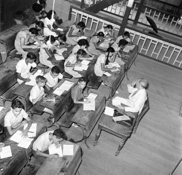 Schwarzes und weißes Foto eines Klassenzimmers mit Kindern an Tischen, Büchern und Gegenständen auf den Tischen und einem Geländer und Schirm im Hintergrund.
