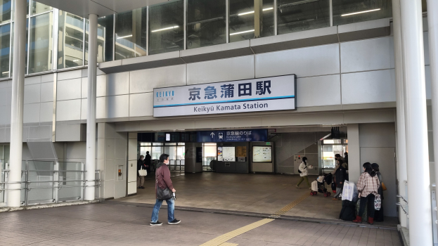 Menschen mit Taschen durch eine Bahnhofshalle gehen, ein Schild mit Text, Säulen, Geländer, Glaswände und Deckenleuchten; ein Mann in Schuhen ist zentral zu sehen.