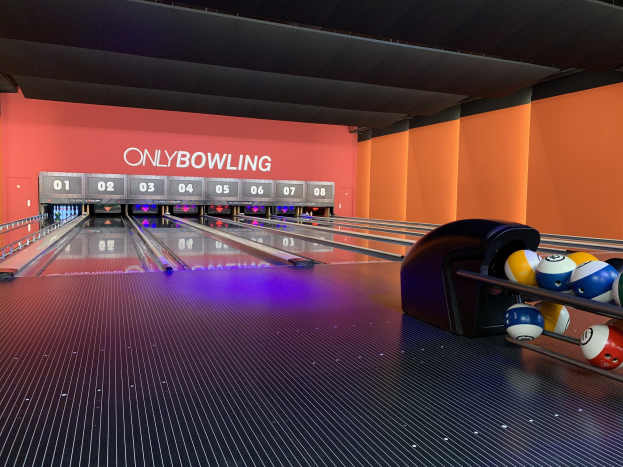 Eine Bowlinganlage mit Bowlingkugeln auf der rechten Seite, Bowlingpins in der Mitte, einer textbedeckten Wand im Hintergrund und einer sichtbaren Decke.