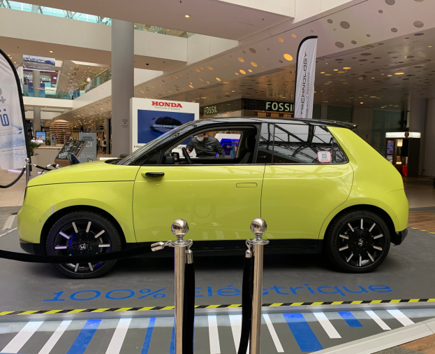 Gelbes wasserstoffbetriebenes Elektroauto vor einem Gebäude mit Absperrpollern, Bannern, Personen, Schildern, einer Topfpflanze, Deckenleuchten und Lüftern.