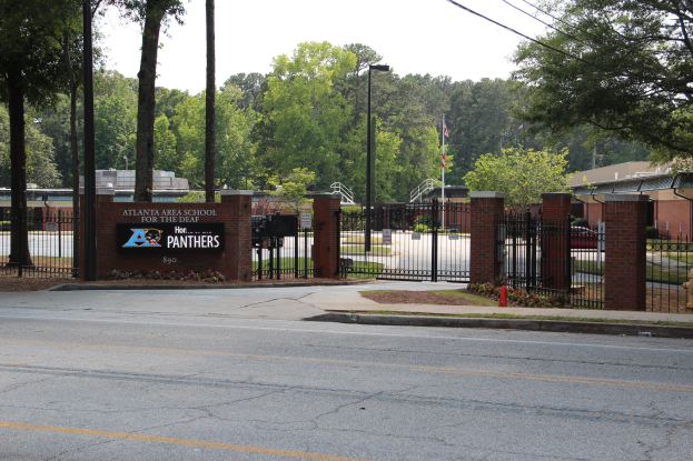 Eintritt in eine Schule mit einem Schild, umgeben von Bäumen, Straßenmöbeln, Fahrzeugen und Gebäuden unter einem klaren blauen Himmel.