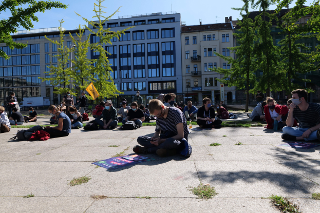 Eine Gruppe von Menschen sitzt vor einem Gebäude auf dem Boden während einer Demonstration in Berlin, einige tragen Masken, mit verstreuten Taschen und Gegenständen, Bäumen und einem klaren blauen Himmel im Hintergrund.