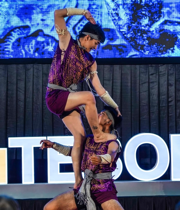 Ein Mann und eine Frau in lila Kleidern f├╝hren Akrobatik auf der B├╝hne aus, wobei der Mann die Frau in seinen Armen h├Ąlt, waehrend eine Menge von unten hinter einem schwarzen Vorhang und einem Bildschirm zusieht.