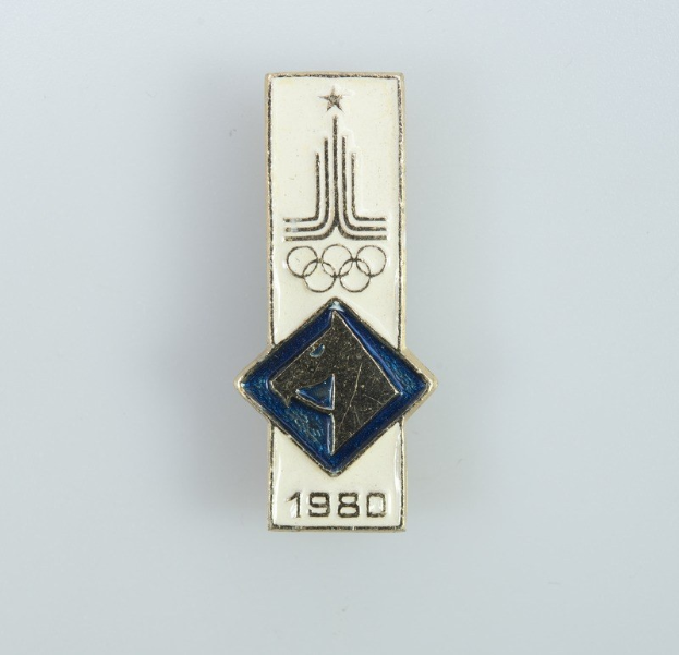 Ein Ansteckabzeichen mit dem Logo der Olympischen Spiele 1980 in Blau und Weiß mit den Olympischen Ringen und der schwarzen Aufschrift "Olympische Spiele 1980".