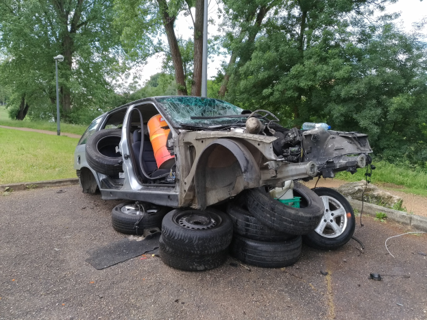 Ein umgekippter Wagen am Straßenrand, umgeben von Gras, Bäumen und Trümmern unter einem bewölkten Himmel.