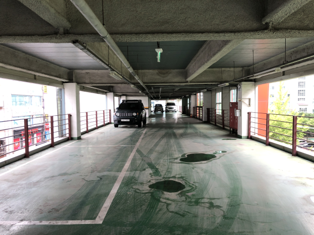 Ein mehrgeschossiges Parkhaus mit parkenden Autos, Geländern, Säulen, Deckenleuchten, freiliegenden Rohren und umliegenden Bäumen vor einem Hintergrund aus Gebäuden mit Fenstern in einer Stadtlandschaft.