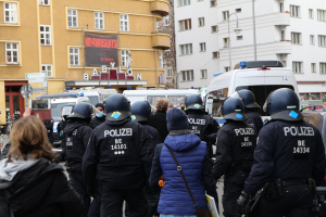 Polizeibeamte in Uniform vor einer Menge bei einer Demonstration in Berlin, mit Fahrzeugen, Gebäuden und einem Kameramann im Bild.