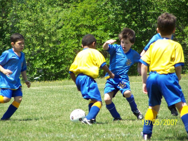 Kinder spielen Fußball auf Rasen mit Bäumen im Hintergrund und einem Datumsstempel in der rechten unteren Ecke.