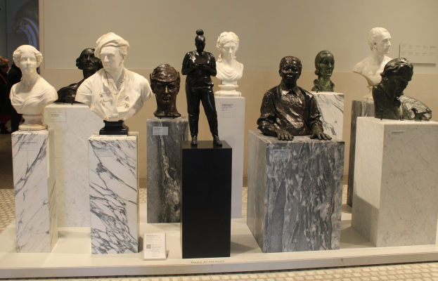 Eine Gruppe detaillierter Büsten-Skulpturen in einem Museum ausgestellt, mit ein paar Menschen im Hintergrund und einer leeren Wand dahinter.