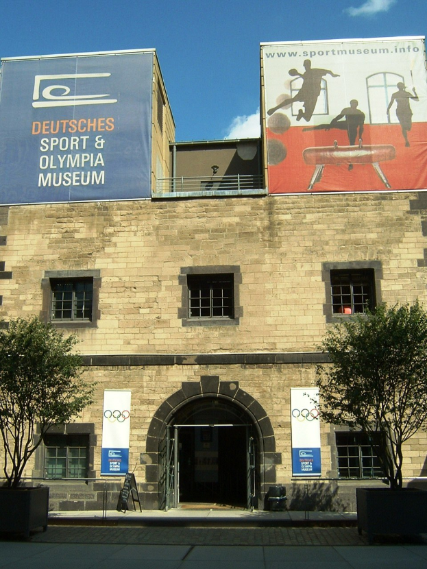 Außenansicht des Deutschen Sport & Olympia Museums in Berlin: Ein Gebäude mit Fenstern und einer Tür, umgeben von Bäumen und Bannern, unter einem bewölkten Himmel.