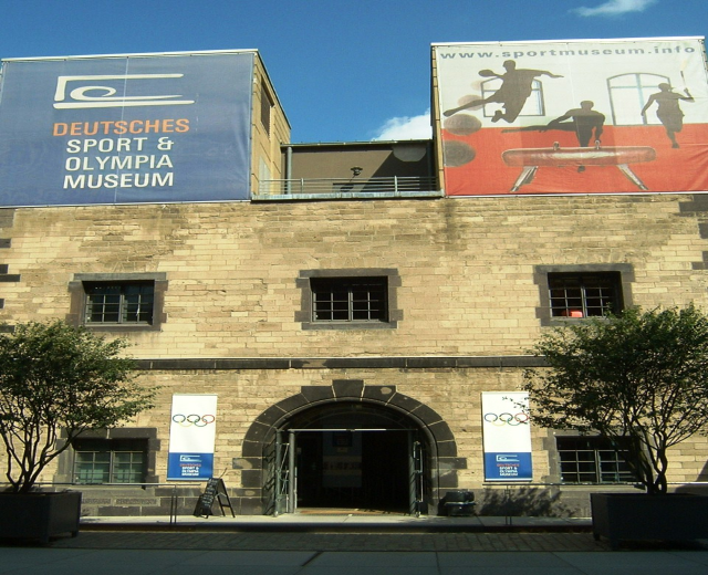 Außenansicht des Deutschen Sport & Olympia Museums in Berlin: Ein Gebäude mit Fenstern und einer Tür, umgeben von Bäumen und Bannern, unter einem bewölkten Himmel.