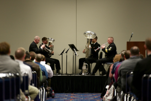 Eine Gruppe von Männern, die auf einer Bühne Blasinstrumente spielen, mit Notenständern, einem Podium und einem Mikrofon, vor einem Publikum, das auf Stühlen sitzt.