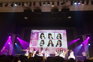 Eine Gruppe von Menschen, die auf einer hell erleuchteten Bühne performen, mit einer Menge darunter und einer Person mit Mikrofon und einer anderen, die ein Instrument spielt, während ein großer Bildschirm Bilder der K-Pop-Gruppe BK48 anzeigt.