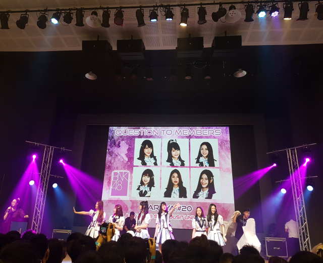 Eine Gruppe von Menschen, die auf einer hell erleuchteten Bühne performen, mit einer Menge darunter und einer Person mit Mikrofon und einer anderen, die ein Instrument spielt, während ein großer Bildschirm Bilder der K-Pop-Gruppe BK48 anzeigt.