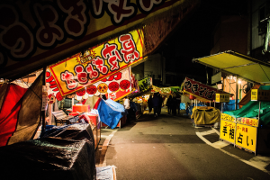 Ein belebter Nachtmarkt mit zahlreichen Ständen und Menschen, flankiert von Gebäuden, beleuchtet von hellen Lichtern, mit einer Person im Vordergrund.