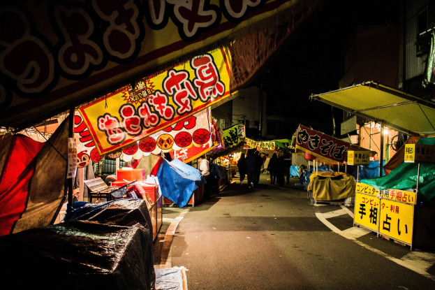 Ein belebter Nachtmarkt mit zahlreichen Ständen und Menschen, flankiert von Gebäuden, beleuchtet von hellen Lichtern, mit einer Person im Vordergrund.