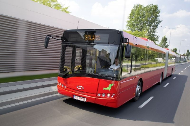 Roter Bus fährt auf einer Straße neben einem Gebäude, mit einer Person im Inneren, neben einer Wand, Bäumen, Laternenpfählen und einem bewölkten Himmel.