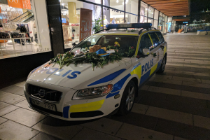 Ein mit Blumen und Blättern geschmückter Polizeiwagen parkt vor einem Geschäft mit Glaswänden bei Nacht, das Schaufensterpuppen in Kleidern und beleuchtete Schilder zeigt, mit Bäumen und Gebäuden im Hintergrund.